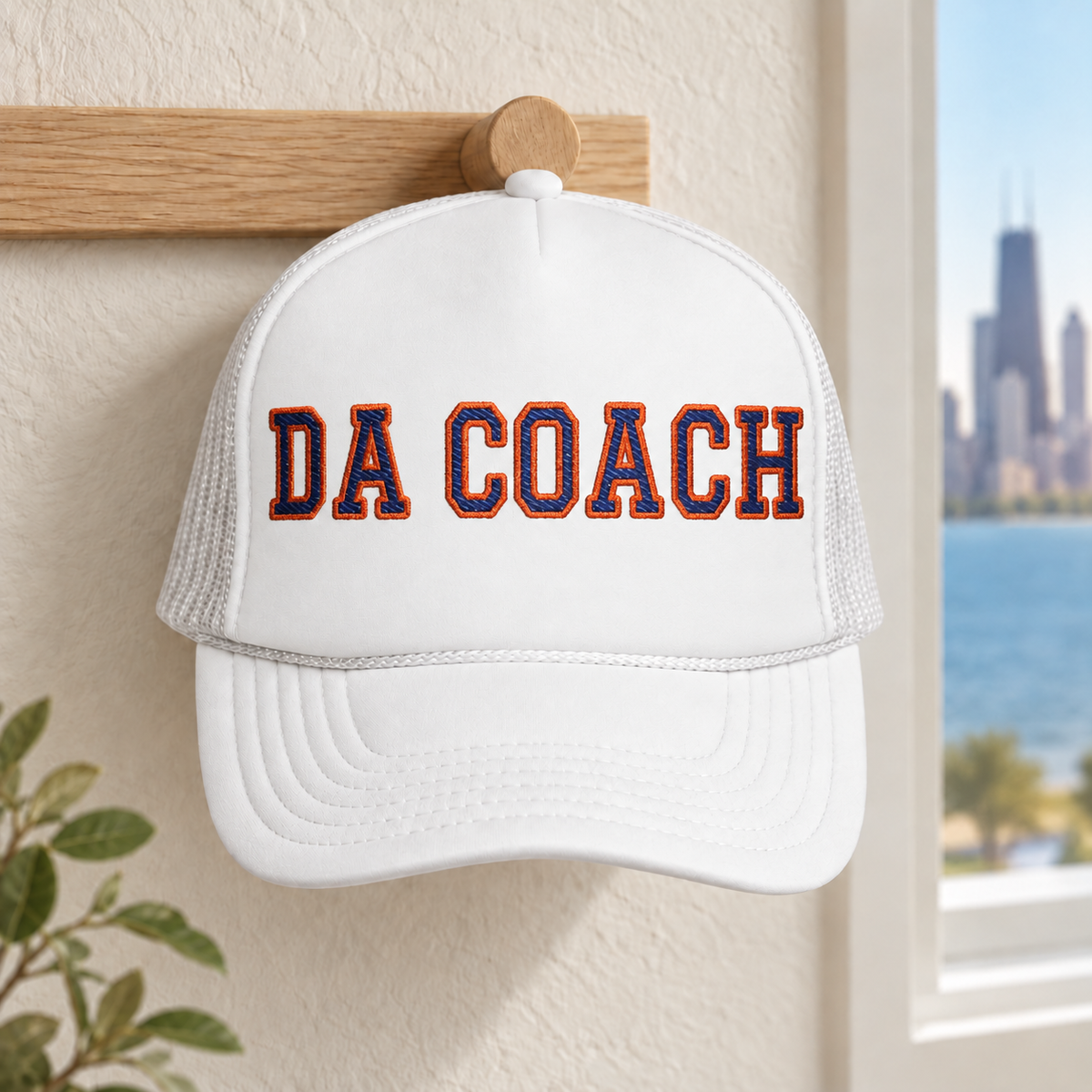 Da Coach embroidered foam trucker hat in all white on a wall hook by a Chicago skyline window