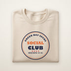 Green Bay Sucks Social Club crewneck in Sand, flat lay with navy and orange circle badge